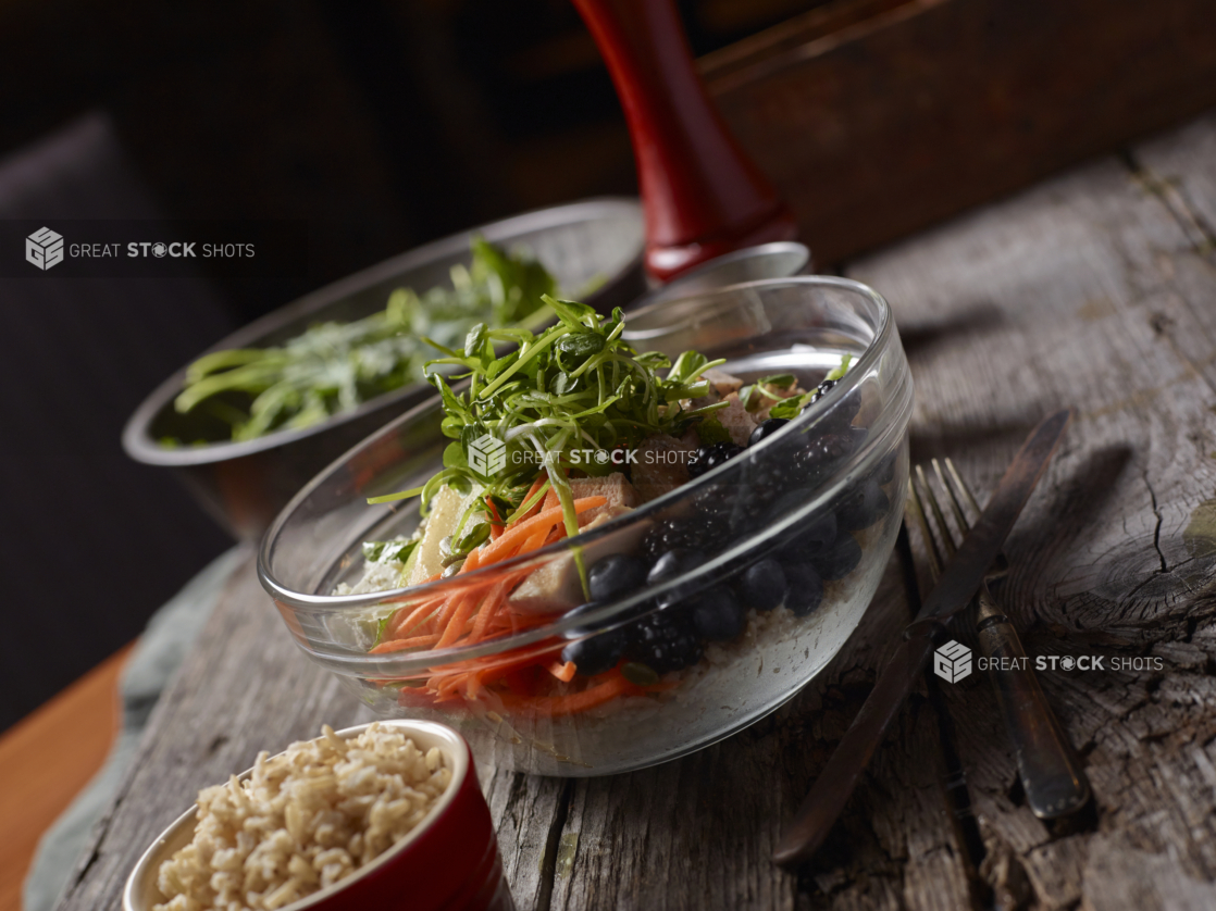 Glass Bowl with Pea Shoots, Julienne Carrots, Grilled Chicken, Tofu and Berries on an Aged Wooden Surface with a Red Ramekin of Brown Rice