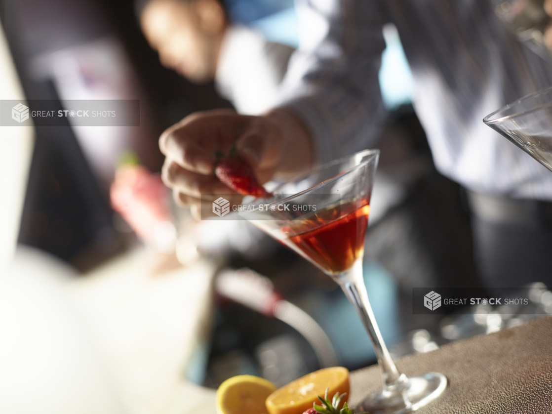 Bartender Adding Strawberry Garnish to a Red Cocktail in a Martini Glass in an Indoor Restaurant Bar Counter Setting