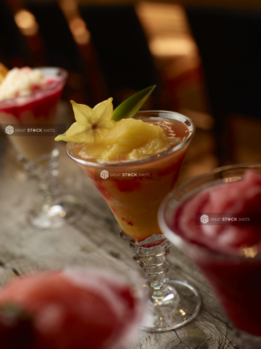 Close Up of a Bellini Cocktail in a Glass Cup with Star Fruit Garnish and other Slushie Cocktails in an Indoor Restaurant Setting