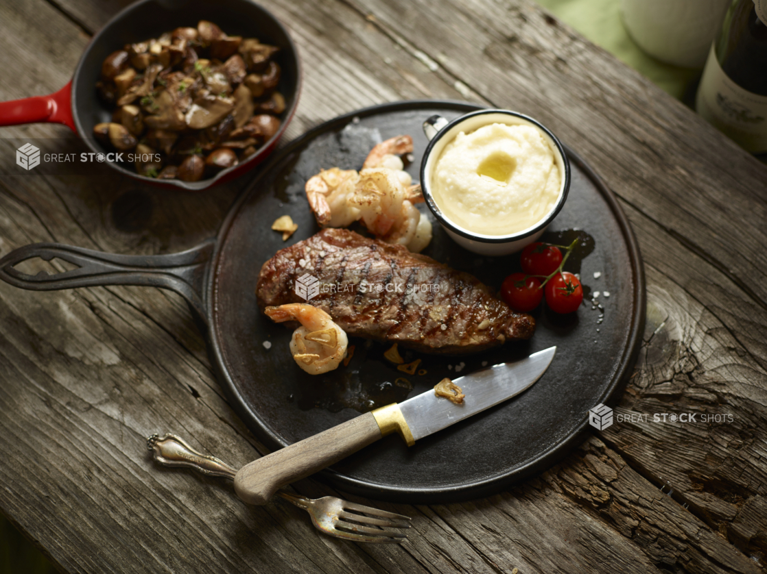 Overhead View of Cast Iron Platter with Surf and Turf Dinner of Sautéed Shrimp and Grilled Sirloin Steak with a Side Bowl of Mashed Potatoes and a Mini Skillet of Sautéed Mushrooms on an Aged Wood Surface