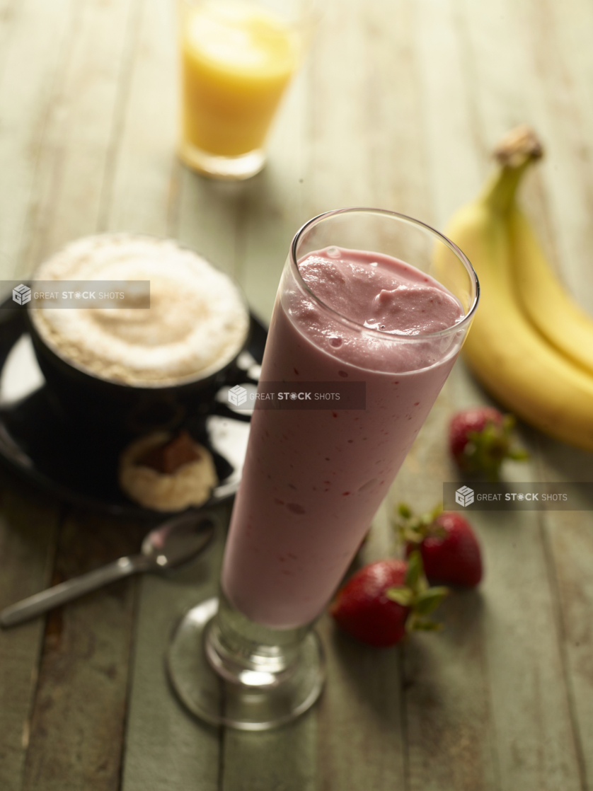 Tall Glass of Strawberry Smoothie on a Painted Weathered Table with Bananas, a Cup of Cappuccino and a Cup of Orange Juice