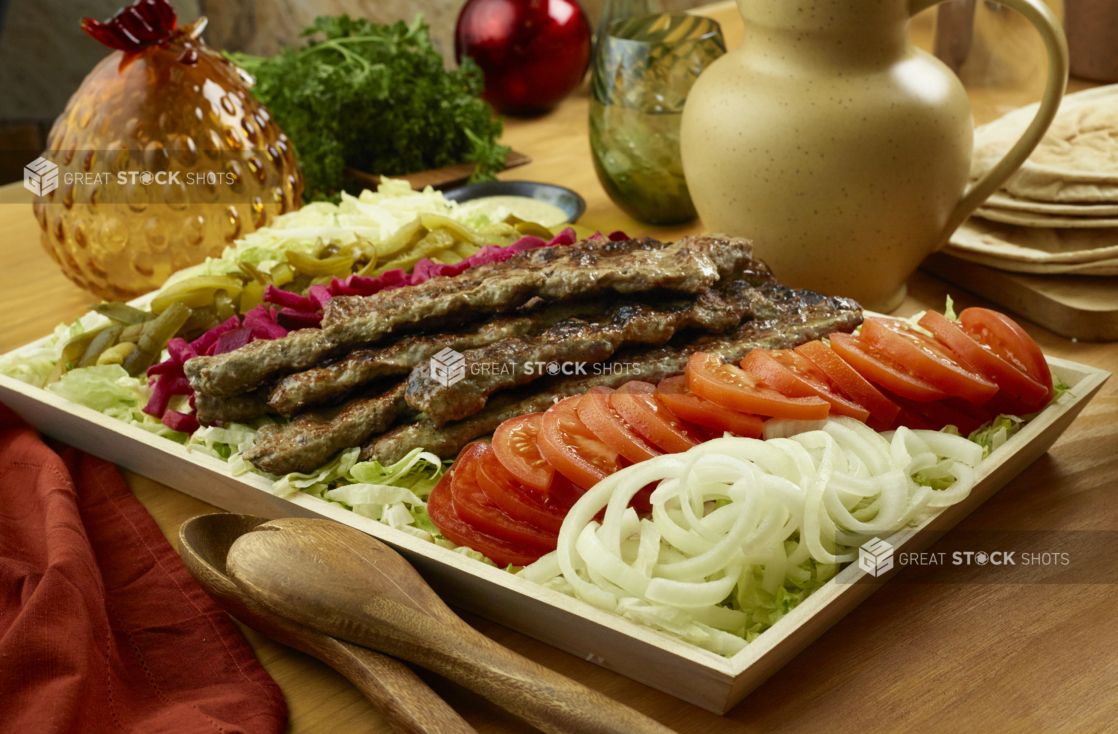 Kebab platter with tomatoes, onion, and pickles on a bed of shredded lettuce on a wood catering tray, wood tabletop, aesthetic objects surrounding