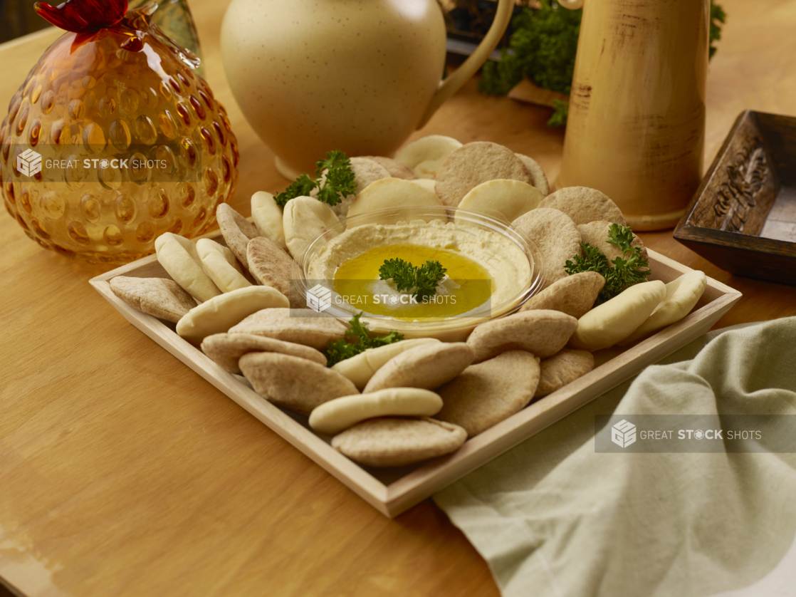 Square Wooden Serving Tray with Hummus Dip and Whole Wheat and White Mini Pita Pockets for Catering on a Wooden Table Surface in an Indoor Setting