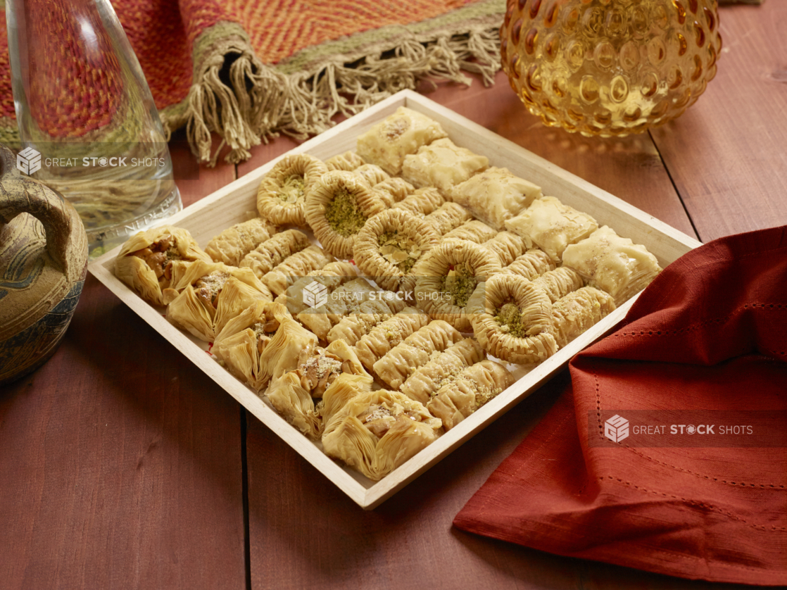 Small Shallow Square Wood Serving Tray of Assorted Baklava for Catering on a Wooden Table with Red Cloth Napkin in an Indoor Setting