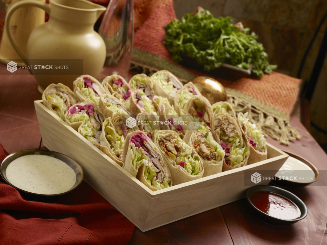 Assorted Middle Eastern Pita Wraps (Chicken and Beef Shawarma, Chicken and Beef Kebabs) in a Deep Square Wood Serving Tray for Catering with Dipping Sauces on a Wooden Table in an Indoor Setting