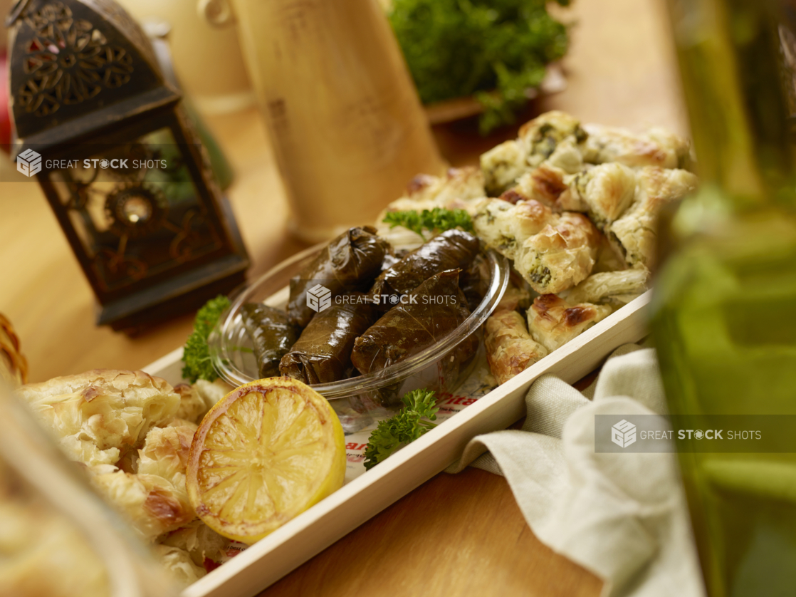 Close Up of Narrow Wood Serving Tray of Middle Eastern Finger Foods Including Marinated Stuff Grape Leaves and Phyllo Pastries on a Wooden Table in an Indoor Setting