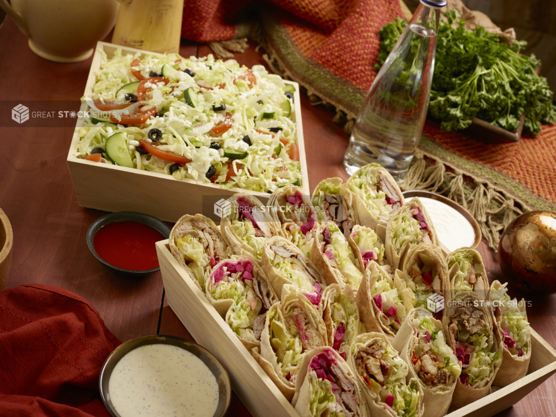 Deep Square Wood Serving Trays with Greek Salad and Assorted Pita Wraps (Shawarma and Kebabs) with Dishes of Dipping Sauce on a Wooden Table in an Indoor Setting