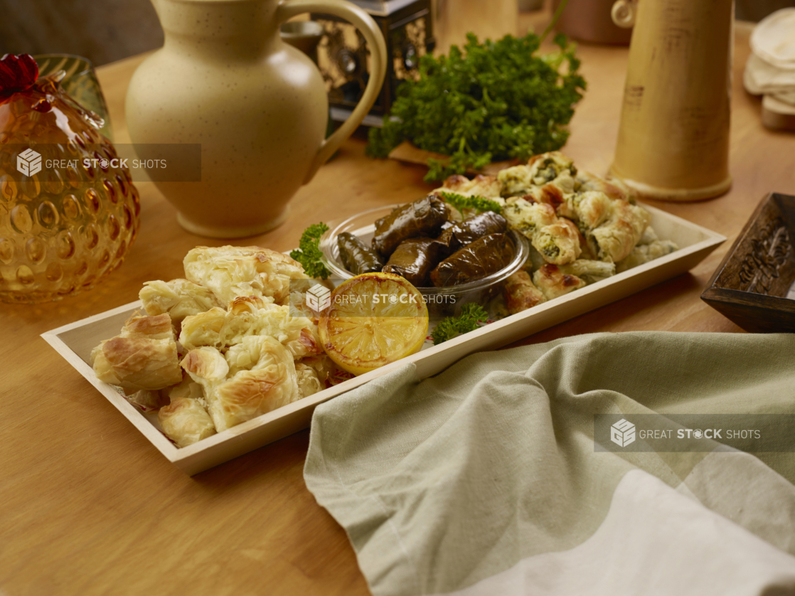 Narrow Wood Serving Tray of Middle Eastern Finger Foods Including Marinated Stuff Grape Leaves and Phyllo Pastries on a Wooden Table in an Indoor Setting