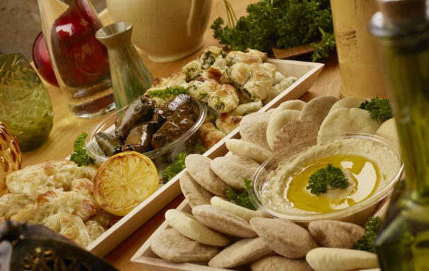 Close Up of Narrow Wood Serving Tray of Middle Eastern Finger Foods and a Square Wood Serving Tray of Hummus and Mini Pitas on a Wooden Table in an Indoor Setting