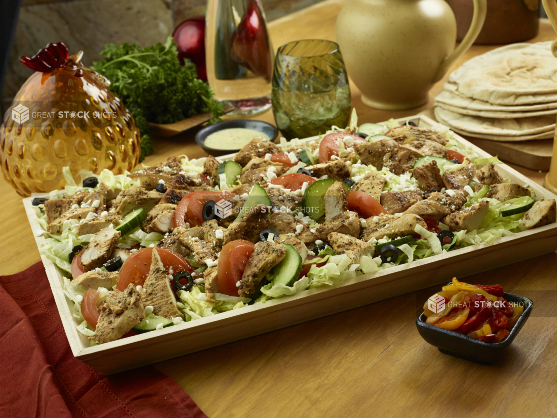 A Large Shallow Wood Serving Tray with a Greek Salad of Sliced Vegetables, Herb-Roasted Chicken Breast, Black Olives and Feta in an Indoor Setting