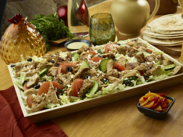A Large Shallow Wood Serving Tray with a Greek Salad of Sliced Vegetables, Herb-Roasted Chicken Breast, Black Olives and Feta in an Indoor Setting