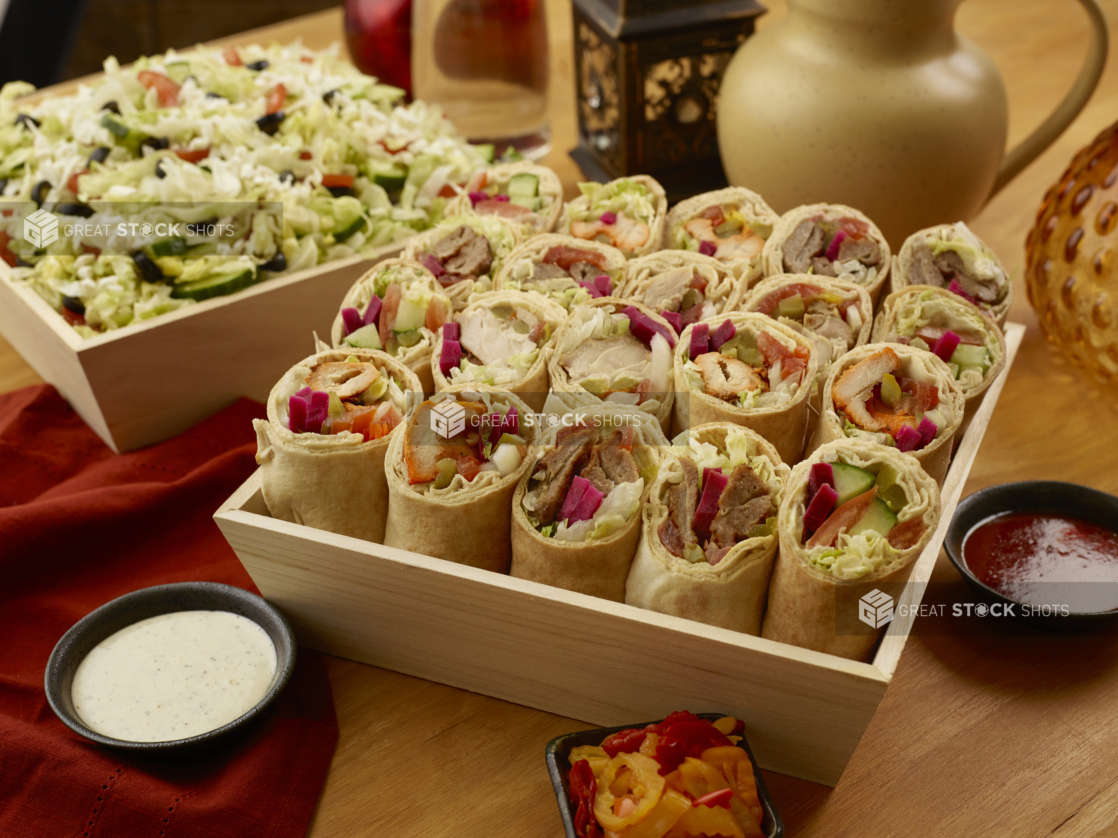 Deep Square Wood Serving Trays with Greek Salad and Assorted Pita Wraps (Shawarma and Kebabs) with Dishes of Dipping Sauce on a Wooden Table in an Indoor Setting - Variation