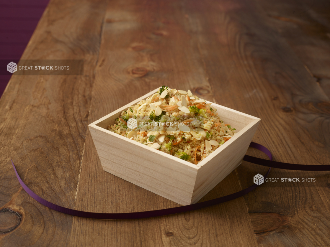 Cous cous salad with sliced almonds, broccoli, and shredded carrots in a wood catering box with a purple ribbon, wood background