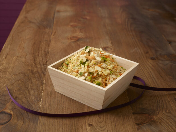 Cous cous salad with sliced almonds, broccoli, and shredded carrots in a wood catering box with a purple ribbon, wood background