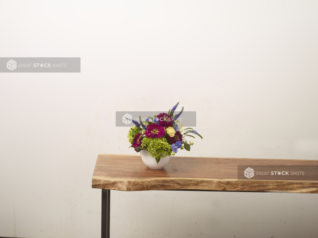 Fuschia, green, and lavender-coloured flower arrangement with zinnias, hydrangeas, and lupines in a white vase positioned at the end of a long wooden table