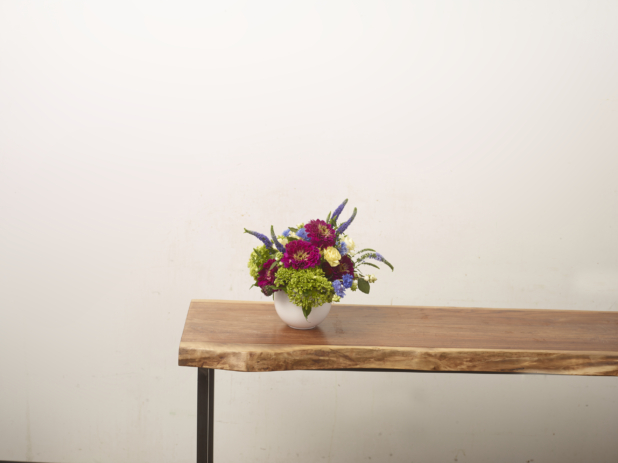 Fuschia, green, and lavender-coloured flower arrangement with zinnias, hydrangeas, and lupines in a white vase positioned at the end of a long wooden table