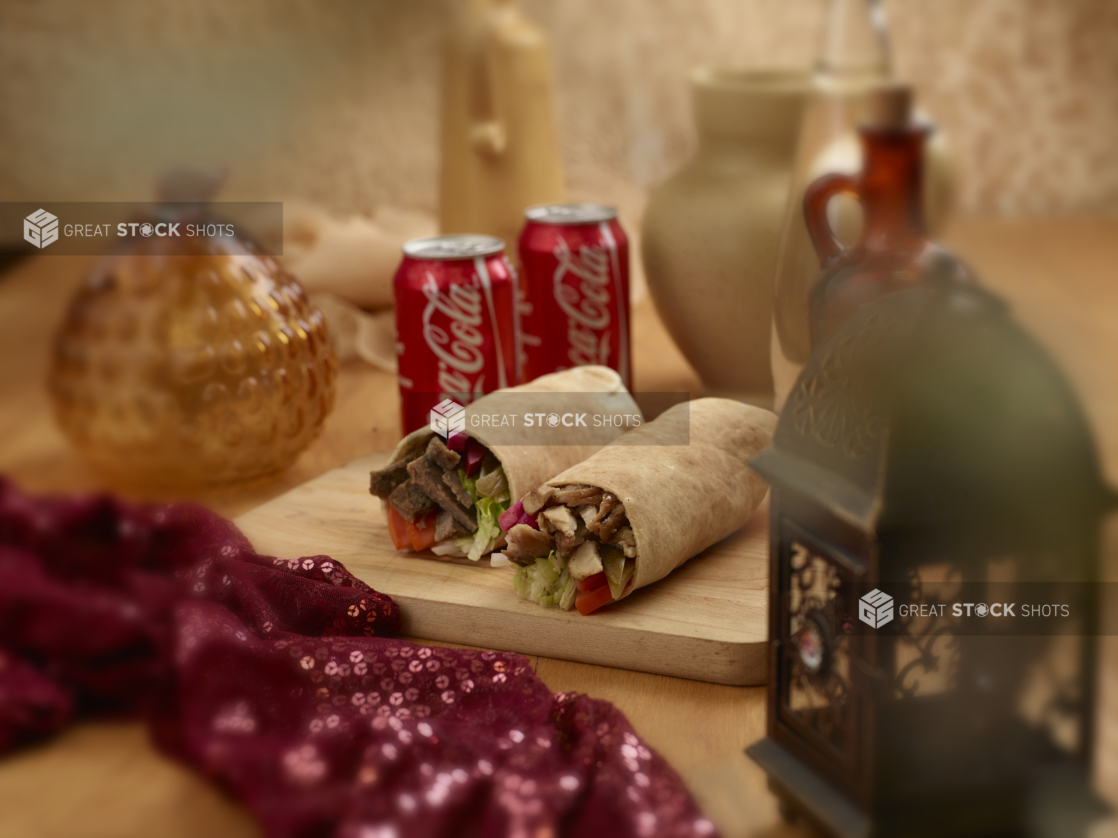 Beef and Chicken Shawarma Pita Wraps with Fresh and Pickled Vegetables on a Wooden Cutting Board with Two Cans of Coca-Cola in an Indoor Setting