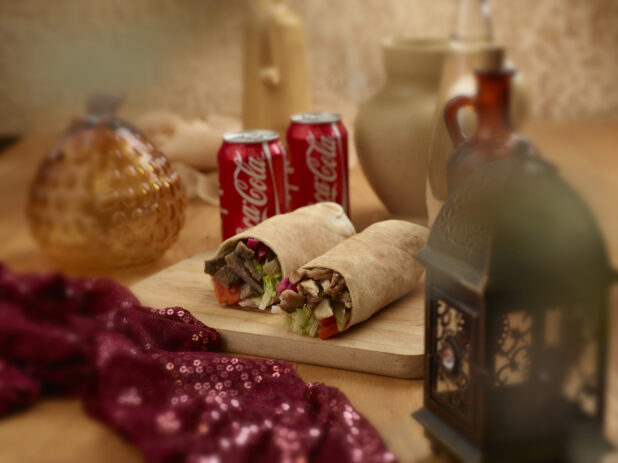 Beef and Chicken Shawarma Pita Wraps with Fresh and Pickled Vegetables on a Wooden Cutting Board with Two Cans of Coca-Cola in an Indoor Setting