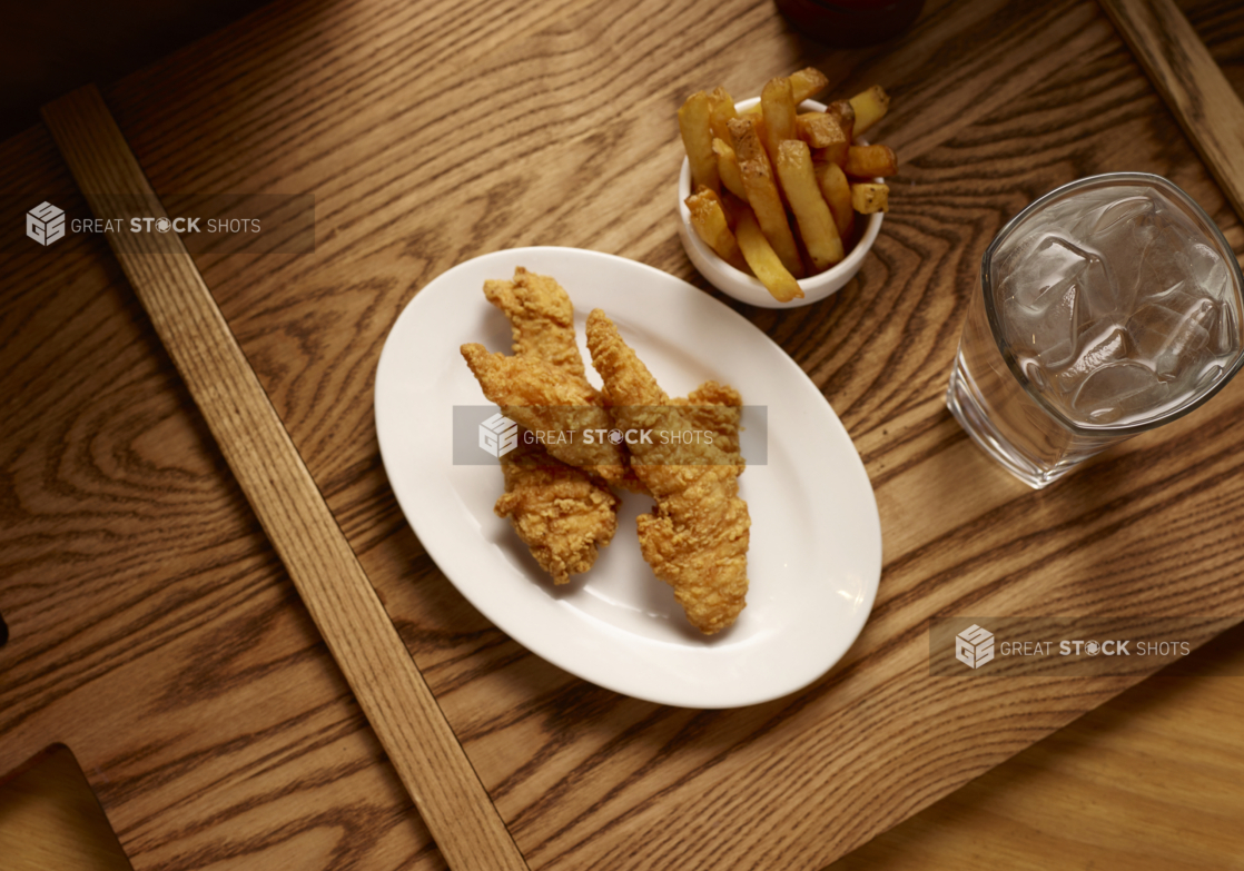 Overhead View of Deep Fried Breaded Chicken Tender Strips on an Oval Plate with a Small White Bowl of Thick Cut Fries with a Glass of Ice Water on a Wooden Cutting Board in an Indoor Setting