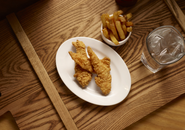 Overhead View of Deep Fried Breaded Chicken Tender Strips on an Oval Plate with a Small White Bowl of Thick Cut Fries with a Glass of Ice Water on a Wooden Cutting Board in an Indoor Setting