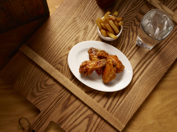 Overhead View of an Oval White Ceramic Dish of Kid-Sized BBQ Chicken Wings and a Small White Bowl of Thick Cut Fries on a Wooden Cutting Board in an Indoor Setting