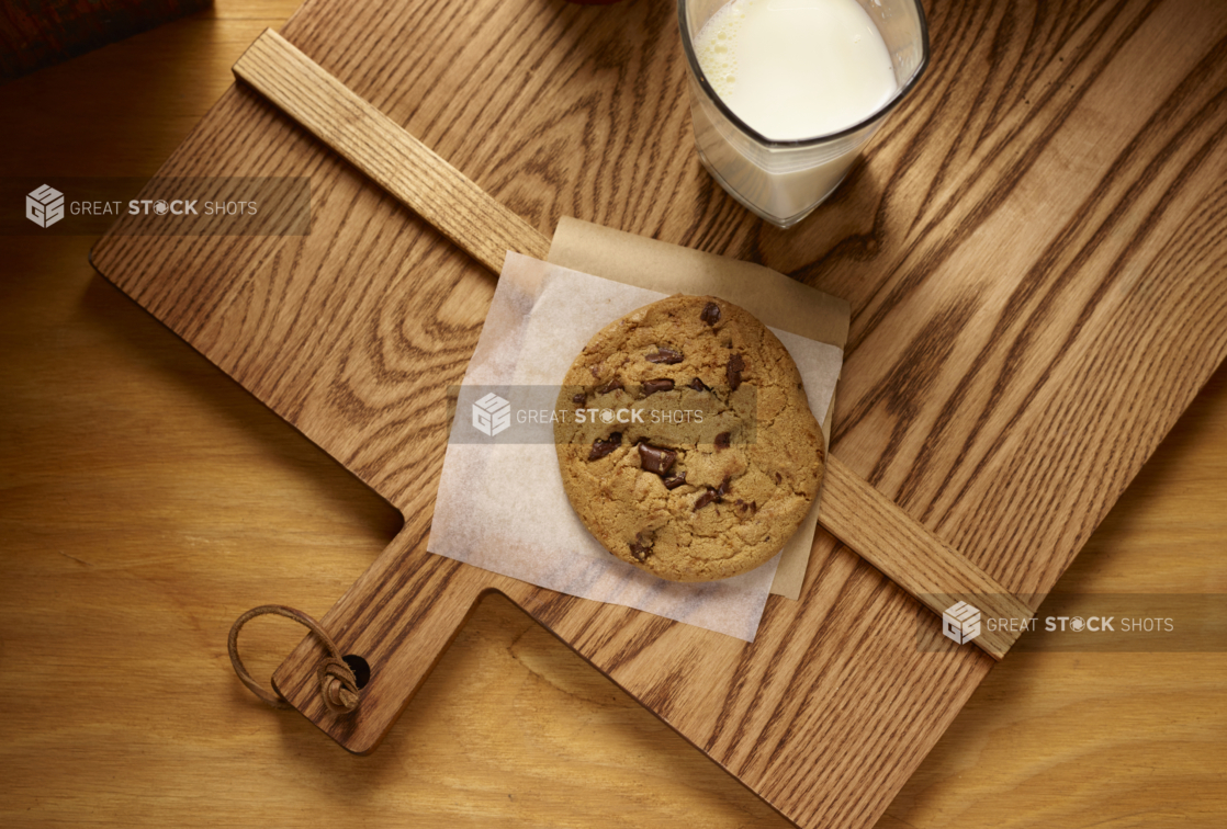 Overhead View of a Jumbo Chocolate Chip Cookie on Parchment Paper with a Large Glass of Milk on a Wooden Cutting Board in an Indoor Setting