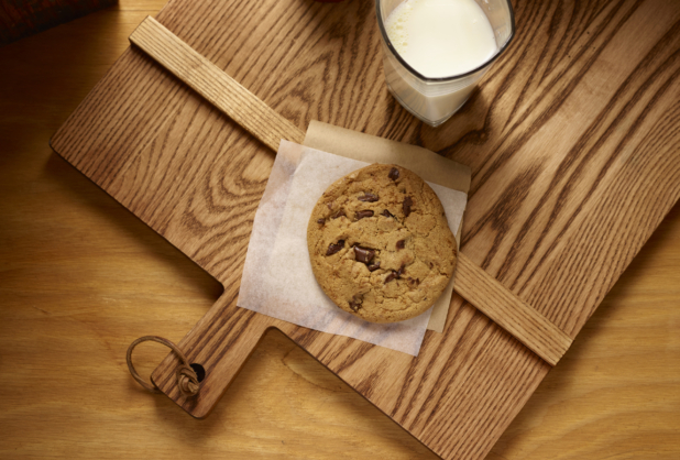 Overhead View of a Jumbo Chocolate Chip Cookie on Parchment Paper with a Large Glass of Milk on a Wooden Cutting Board in an Indoor Setting