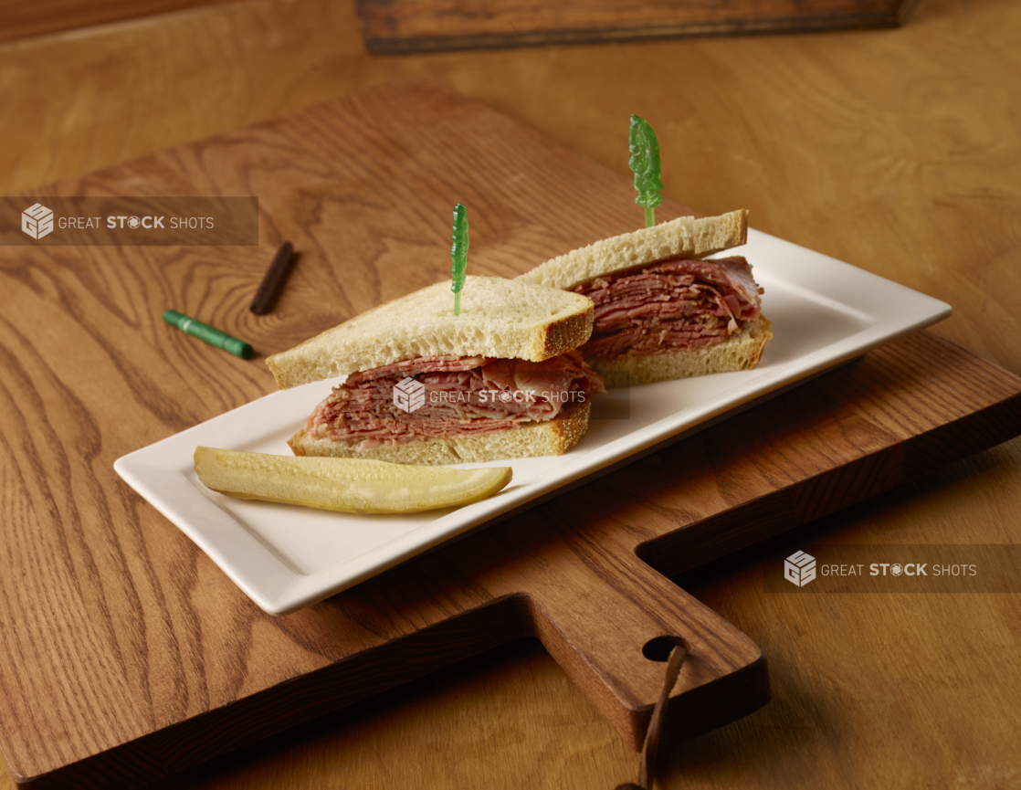 Kids Corned Beef Sandwich on White Rye Bread with a Dill Pickle Spear on a Rectangular White Ceramic Platter on a Wooden Cutting Board in an Indoor Setting