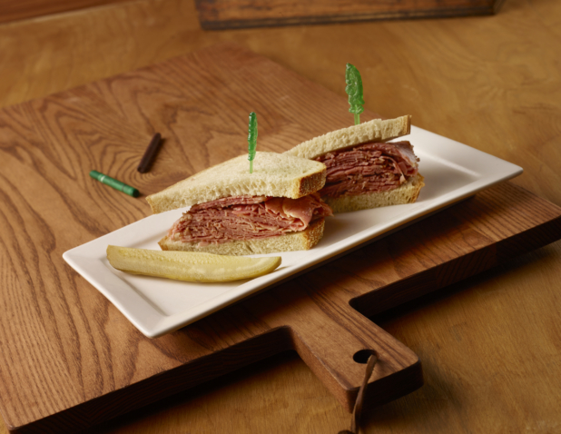 Kids Corned Beef Sandwich on White Rye Bread with a Dill Pickle Spear on a Rectangular White Ceramic Platter on a Wooden Cutting Board in an Indoor Setting