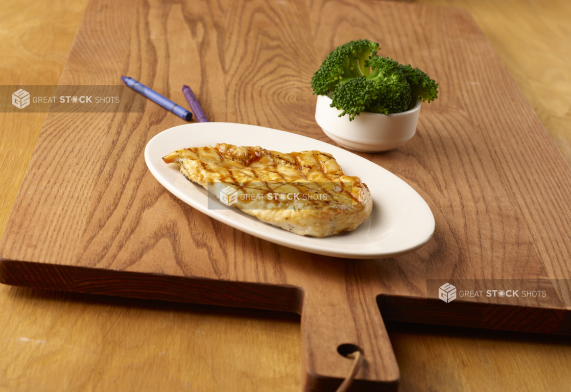 Kids Meal of Grilled BBQ Chicken Breast on a Round White Dish and a Kid-Sized Steamed Broccoli Side Dish on a Wooden Cutting Board in an Indoor Setting