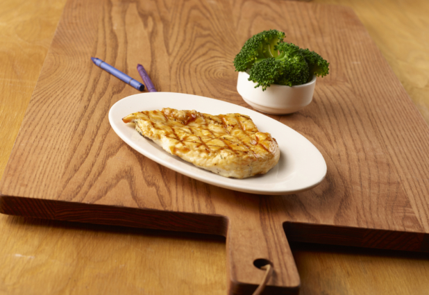 Kids Meal of Grilled BBQ Chicken Breast on a Round White Dish and a Kid-Sized Steamed Broccoli Side Dish on a Wooden Cutting Board in an Indoor Setting