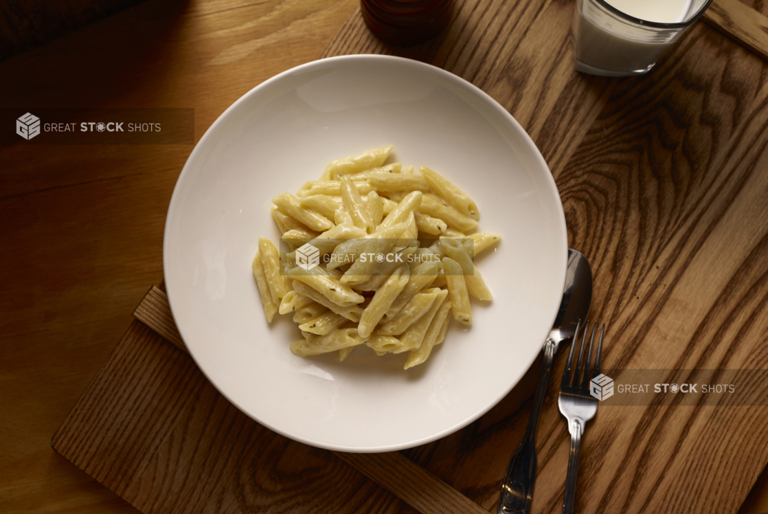 Overhead View of a Kids Penne Alfredo Pasta in a Large White Ceramic Bowl on a Wooden Cutting Board in an Indoor Setting