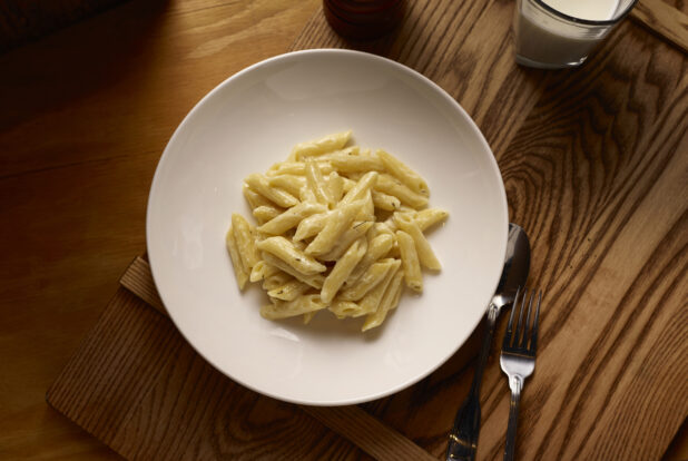 Overhead View of a Kids Penne Alfredo Pasta in a Large White Ceramic Bowl on a Wooden Cutting Board in an Indoor Setting