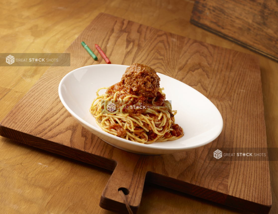 Kids Spaghetti and Meatball in a Large White Ceramic Bowl on a Wooden Cutting Board in an Indoor Setting