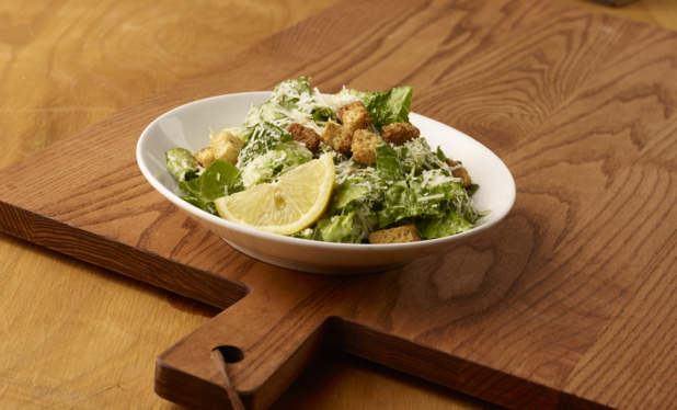 Small Caesar Salad with a Lemon Wedge in a Round White Ceramic Dish on a Wooden Cutting Board in an Indoor Setting