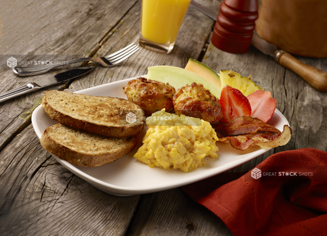 Breakfast Platter of Scrambled Eggs, Bacon, Fresh Fruit, Potato Latkes and Toasted Rye Bread on a Rectangular White Ceramic Platter on a Rustic Wood Surface