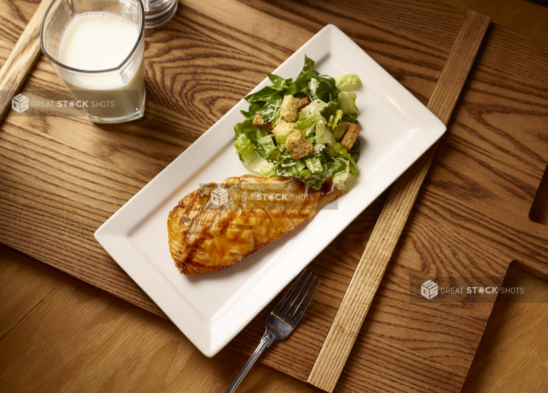 Overhead View of a Kids Grilled Salmon Teriyaki and Caesar Salad Combo Meal on a Rectangular White Ceramic Platter on a Wooden Cutting Board in an Indoor Setting