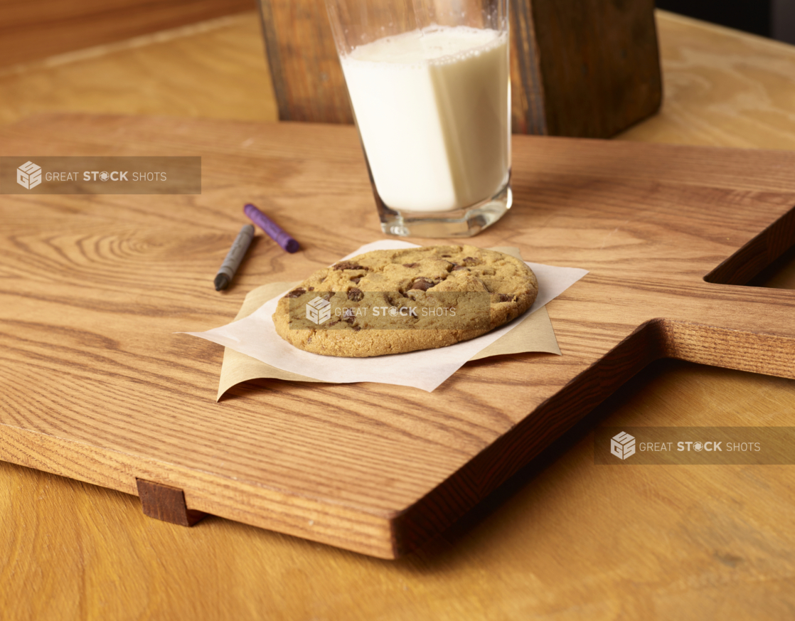 Jumbo Chocolate Chip Cookie on Parchment Paper with a Large Glass of Milk on a Wooden Cutting Board in an Indoor Setting