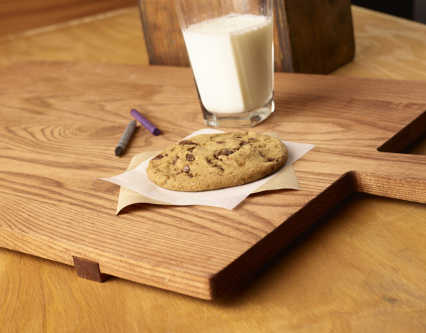 Jumbo Chocolate Chip Cookie on Parchment Paper with a Large Glass of Milk on a Wooden Cutting Board in an Indoor Setting