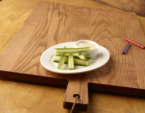 A Round White Dish of Cucumber Sticks and Creamy Ranch Dip in a Ramekin on a Wooden Cutting Board with Crayons for Kids
