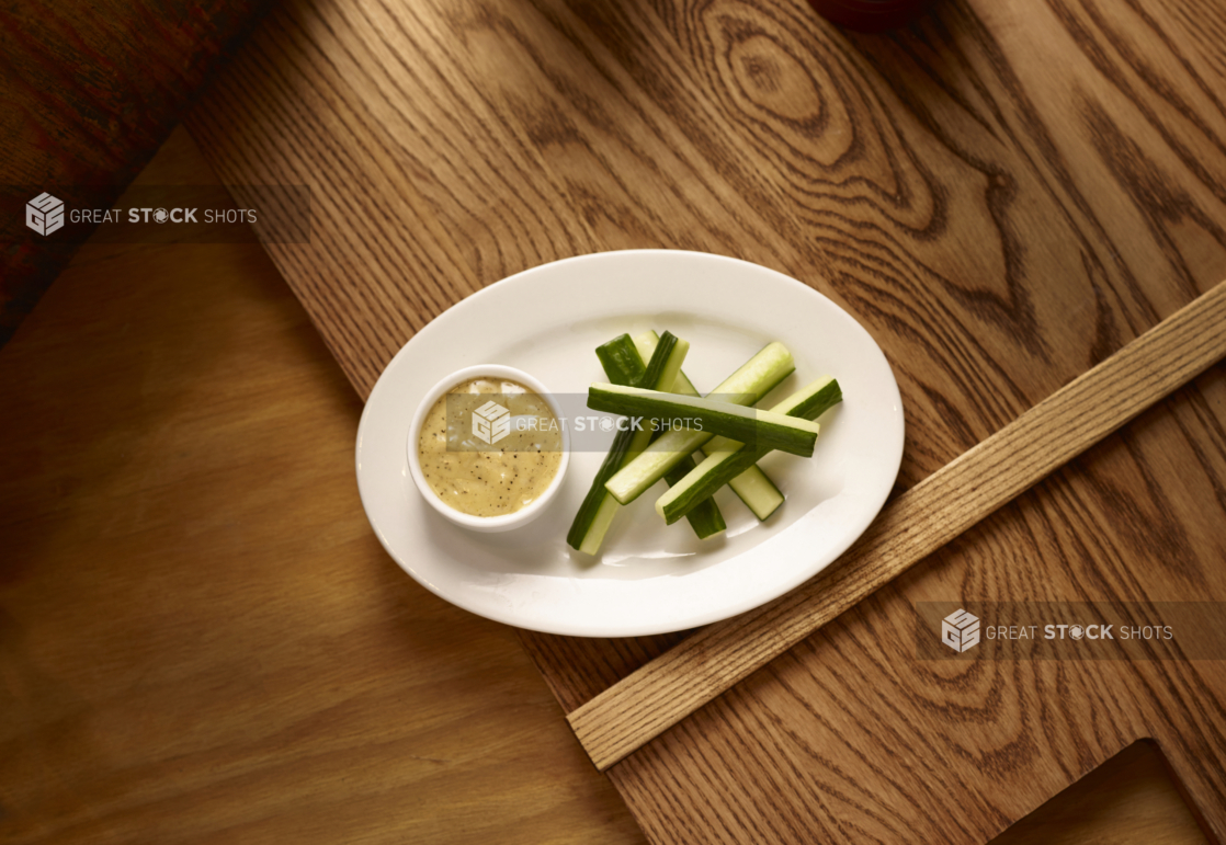 An Overhead View of an Oval White Dish of Cucumber Sticks and Creamy Ranch Dip in a Ramekin on a Wooden Cutting Board in an Indoor Setting