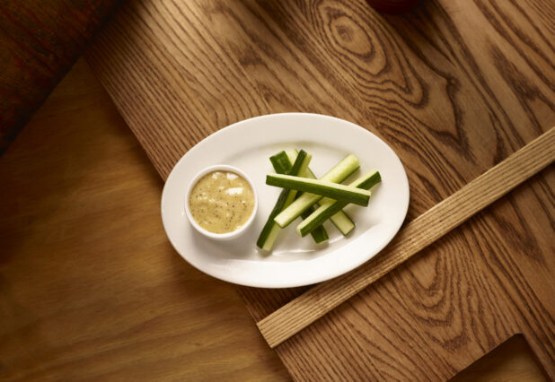 An Overhead View of an Oval White Dish of Cucumber Sticks and Creamy Ranch Dip in a Ramekin on a Wooden Cutting Board in an Indoor Setting