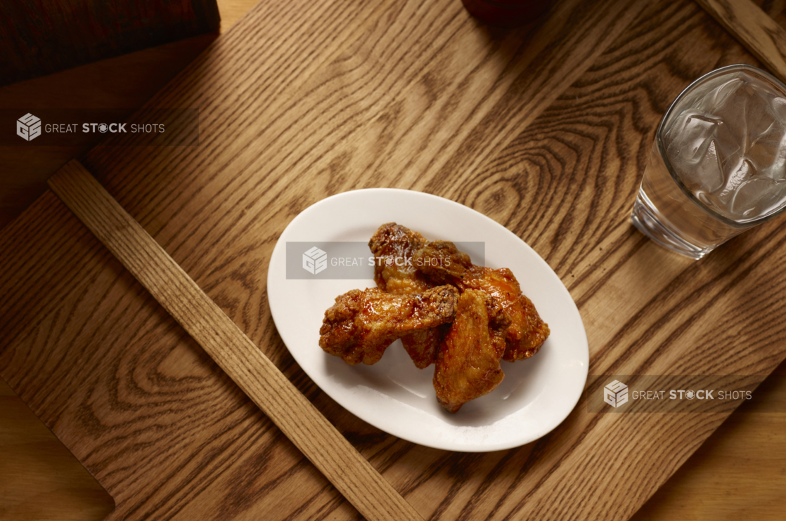 Overhead View of an Oval White Ceramic Dish of Kid-Sized BBQ Chicken Wings and a Glass of Ice Water on a Wooden Cutting Board in an Indoor Setting