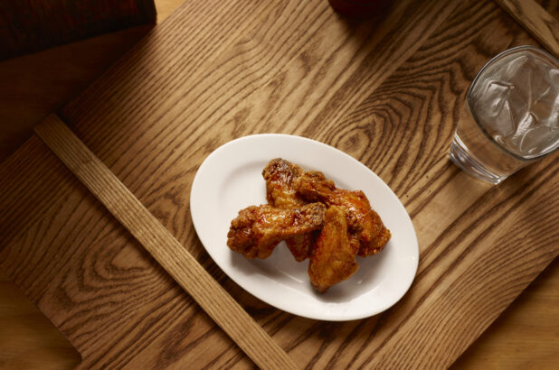 Overhead View of an Oval White Ceramic Dish of Kid-Sized BBQ Chicken Wings and a Glass of Ice Water on a Wooden Cutting Board in an Indoor Setting