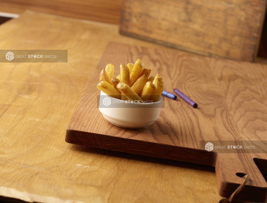 Small Kids Size Thick Cut French Fries in a Round White Bowl on a Wooden Cutting Board on a Wood Surface with Crayons for Kids