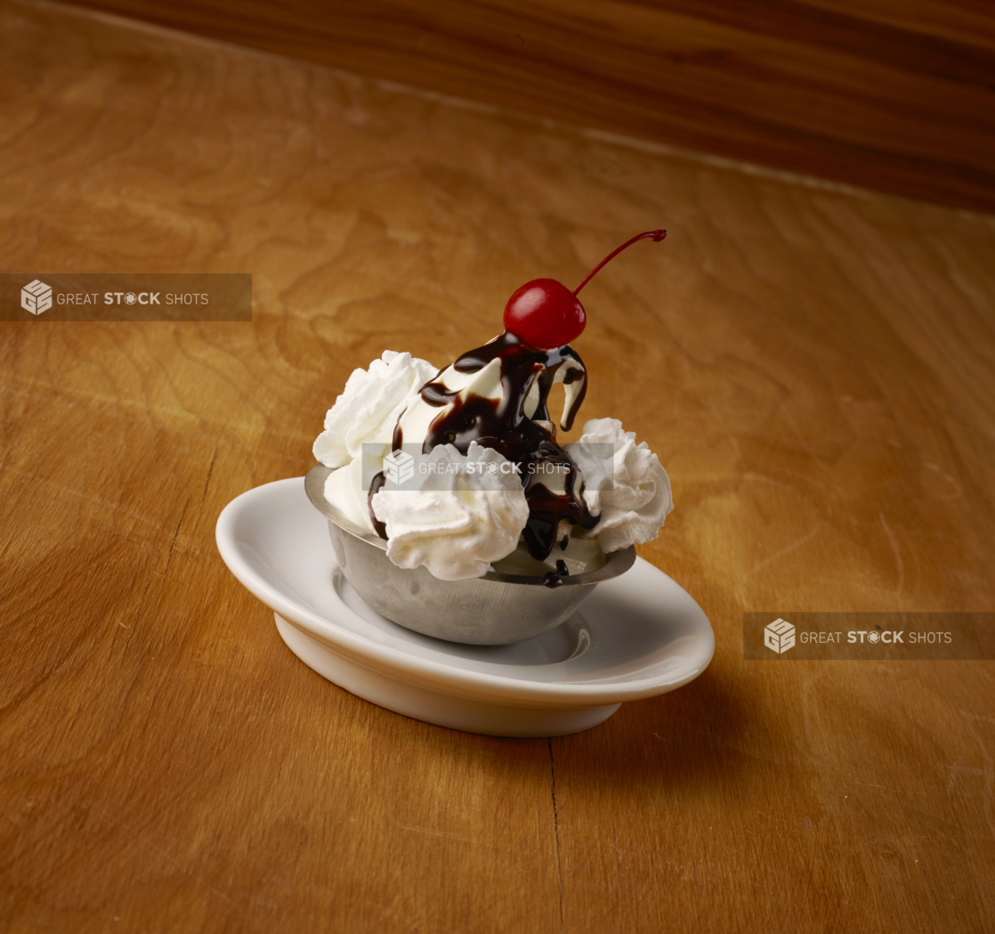 Vanilla Ice Cream Sundae with Chocolate Sauce, Maraschino Cherry and Whipped Cream in a Stainless Steel Bowl on a Wooden Table