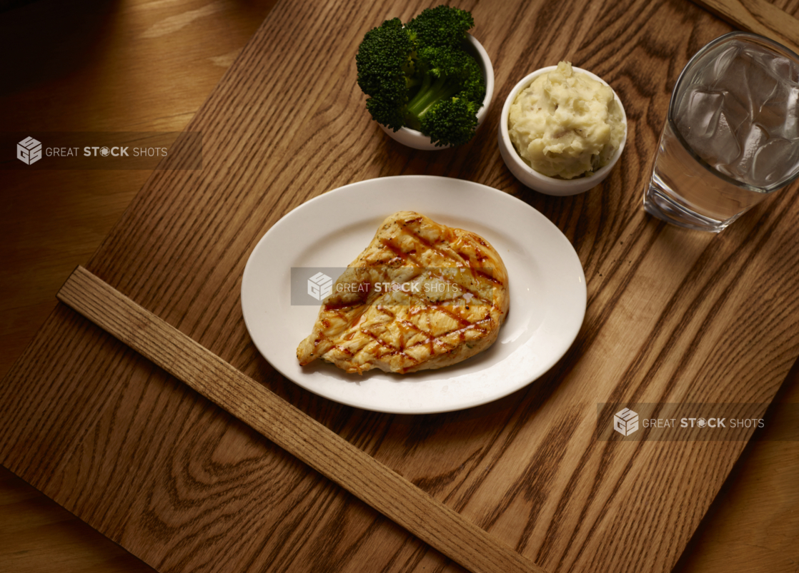 Overhead View of a Kids Meal of Grilled BBQ Chicken Breast, Kid-Sized Steamed Broccoli and Mashed Potato Side Dishes on a Wooden Cutting Board in an Indoor Setting