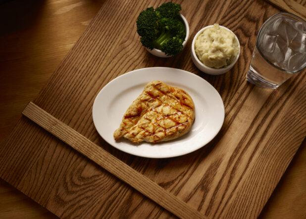 Overhead View of a Kids Meal of Grilled BBQ Chicken Breast, Kid-Sized Steamed Broccoli and Mashed Potato Side Dishes on a Wooden Cutting Board in an Indoor Setting