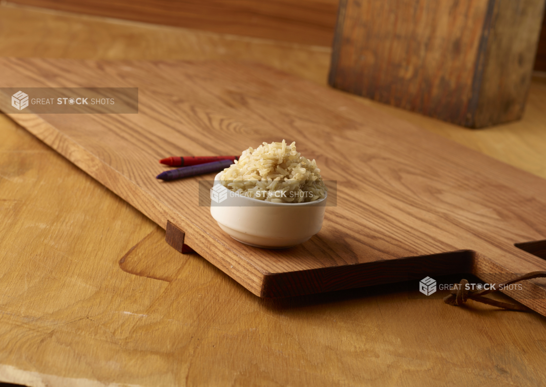 Small Kids Size Brown Rice in a Round White Bowl on a Wooden Cutting Board on a Wood Surface with Crayons for Kids