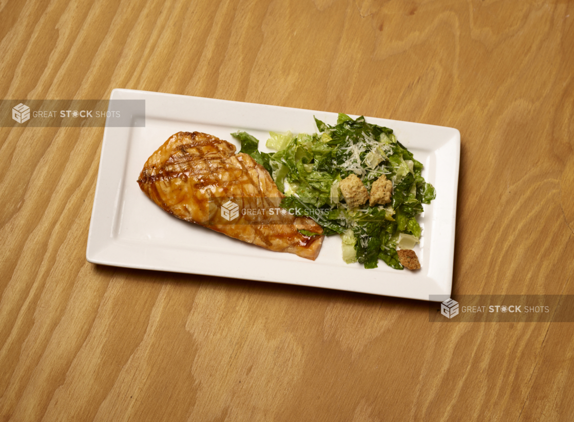 Overhead View of Grilled Salmon Teriyaki with a Side of Caesar Salad on a White Rectangular Platter Dish on a Wooden Surface